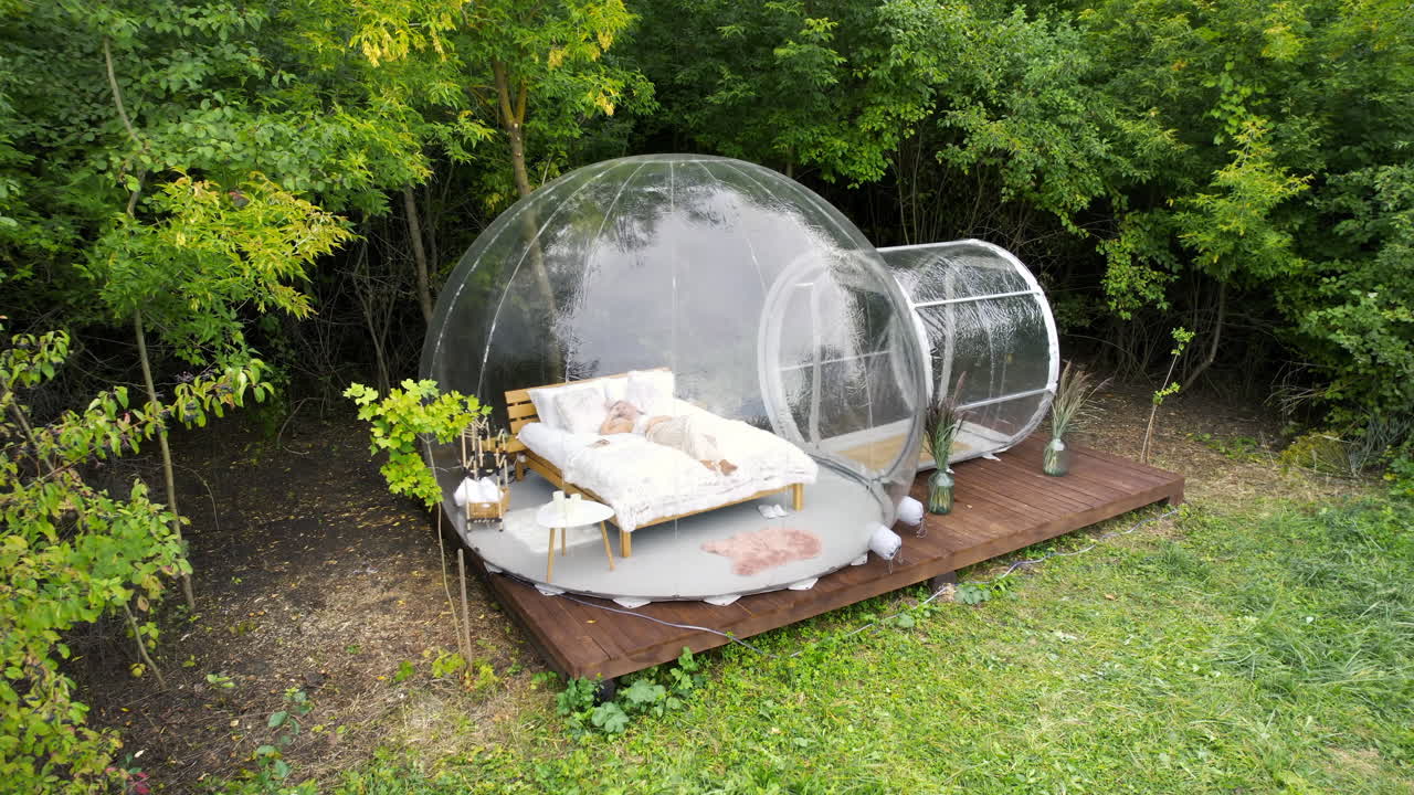 Gennemsigtigt bobletelt ved glamping, frodig skov rundt om og indeni. Kvinde ligger på sengen