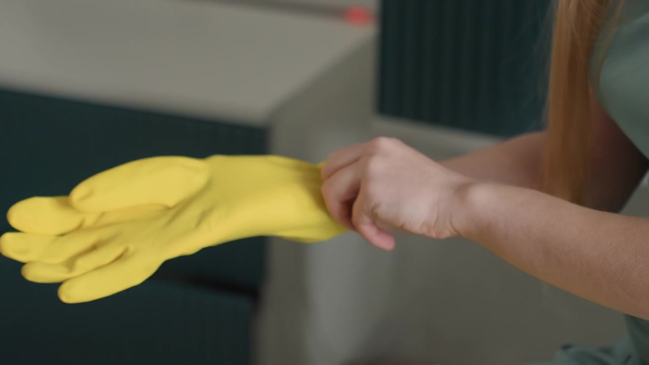 White woman putting on yellow gloves, sitting in living room, sliding rubber gloves over fingers, textured grip visible, calm methodical movements, soft natural light, vintage dress, closeup household
