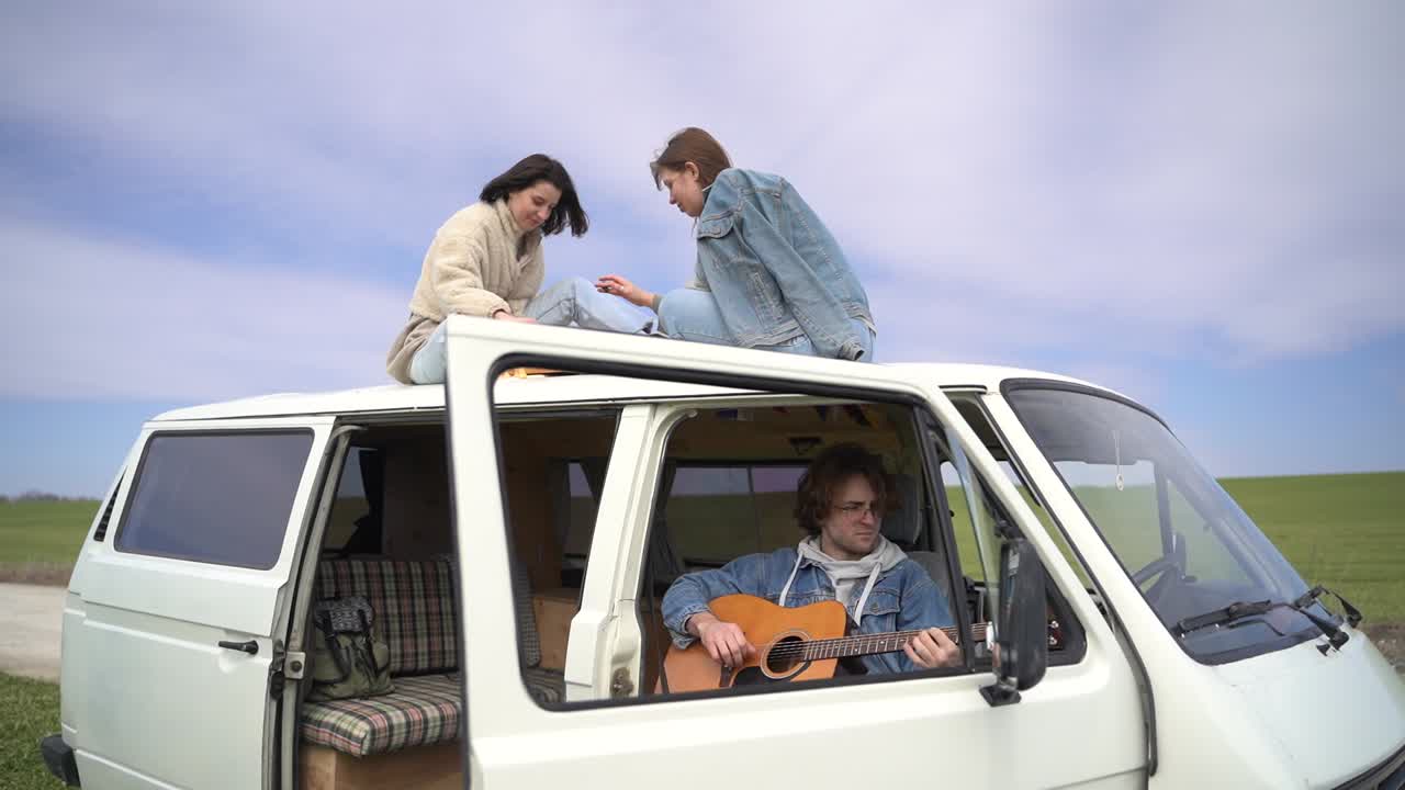 две молодые девушки играют в шахматы на крыше автофургона на дороге, затерянной среди сельской местности. молодой парень играет на гитаре.