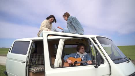 two young girls play chess on the roof of a caravan on a road lost in the middle of the countryside. a young guy plays the guitar.