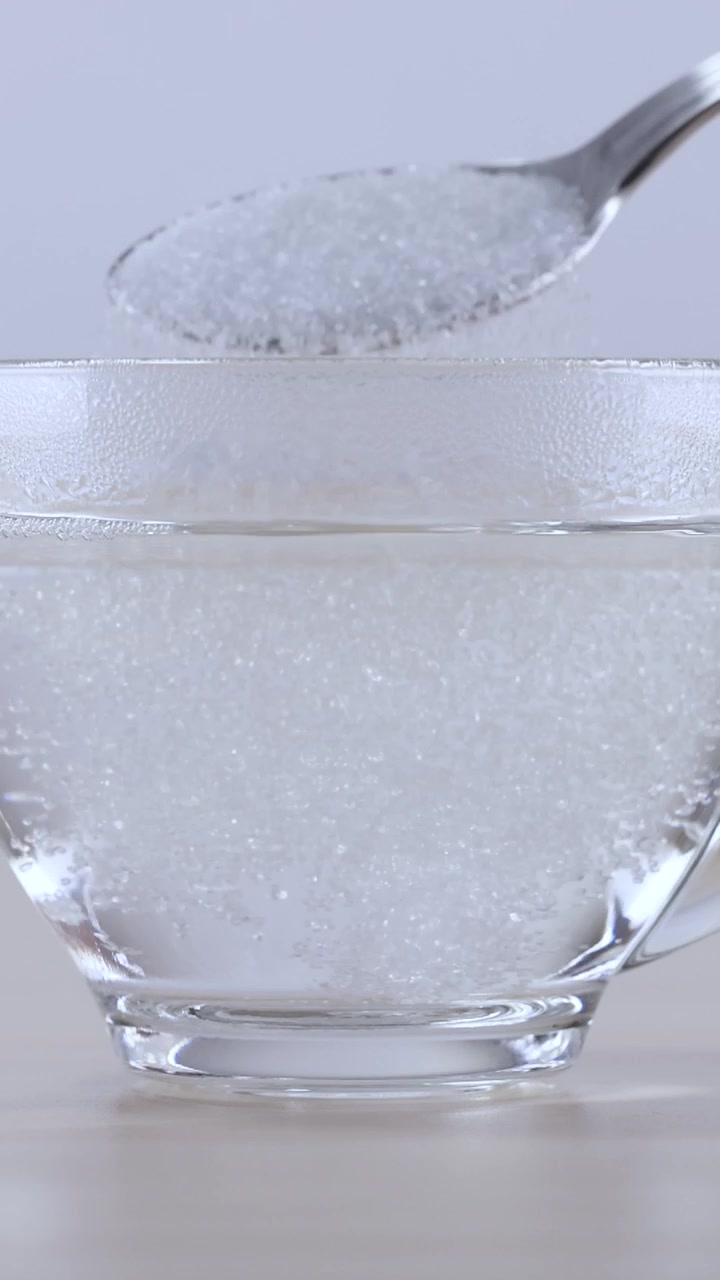 Primo piano di cristalli di zucchero bianco che cadono nell'acqua e si dissolvono in un bicchiere di vetro.