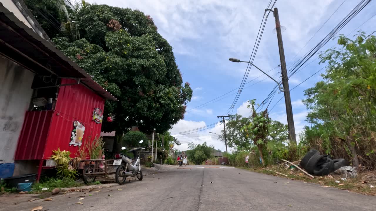 Camera moves along quiet rural road with houses, greenery, and overcast sky in tropical Thailand