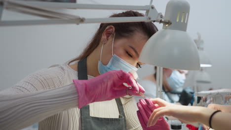 nail technician wearing face mask and pink gloves carefully using scissors and brush on customer nails under bright salon lamp with blurred support arm and second technician visible in background