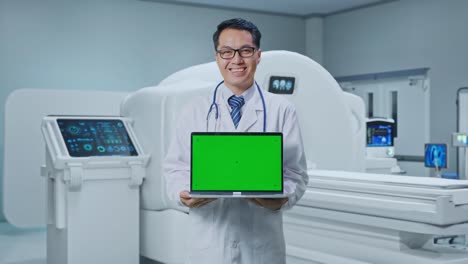 asian male doctor smiling and showing green screen laptop while standing with mri machine in the hospital