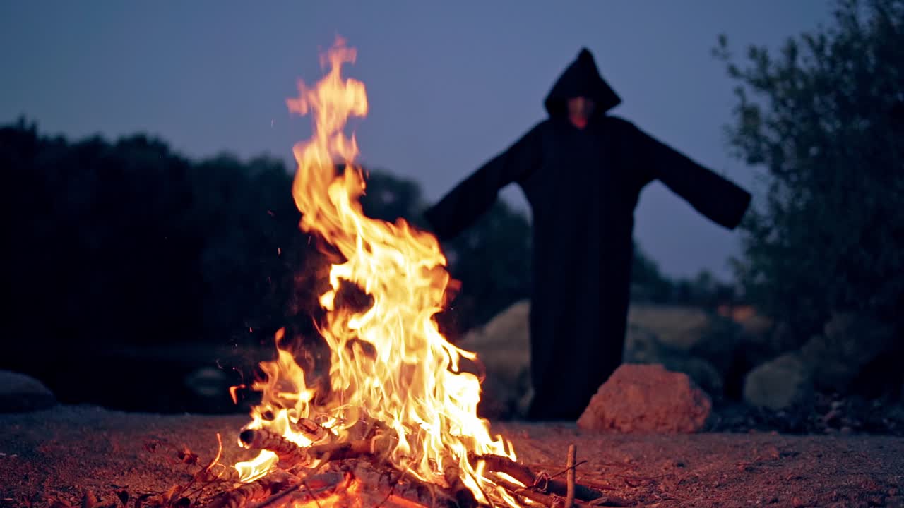 Silhouette of black death by the fire at night with hands outstretched. Freedom concept. Flame on the ground and witch costume on the natural background in the evening.