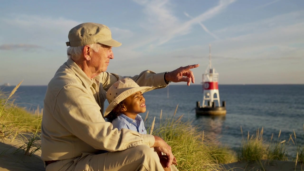 Anziano che indica con il braccio teso, mostrando un faro rosso e bianco a un bambino con il cappello su una duna.