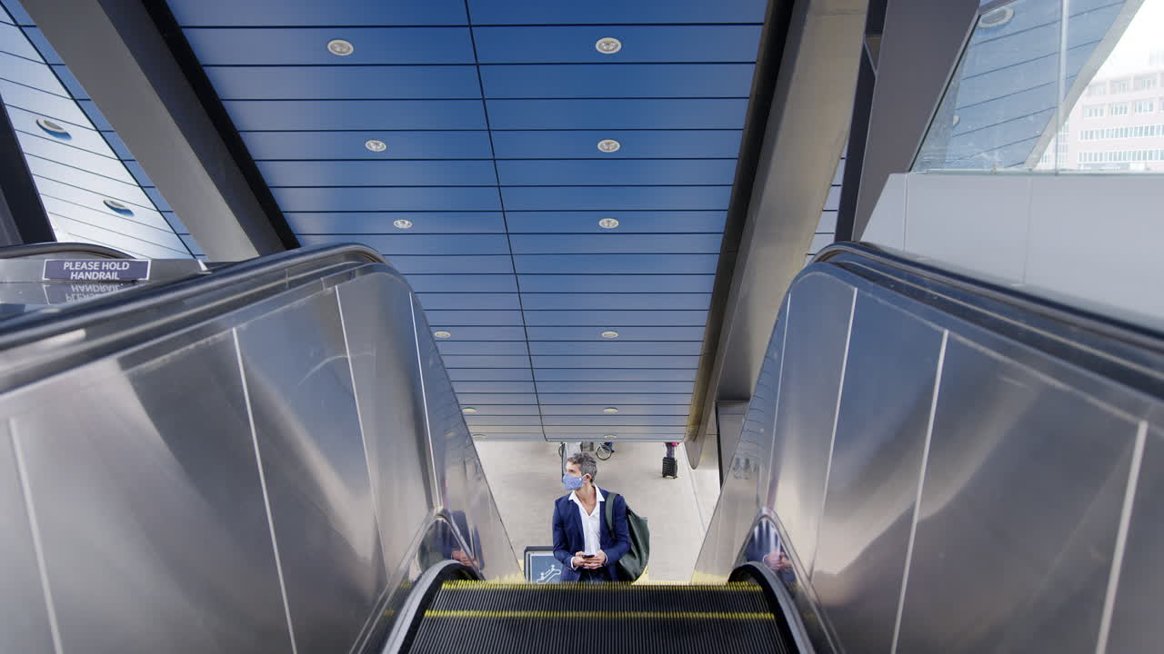 Forretningsmand på rulletrappe på togstation med mobiltelefon, bærer ansigtsmaske (PPE) under pandemi
