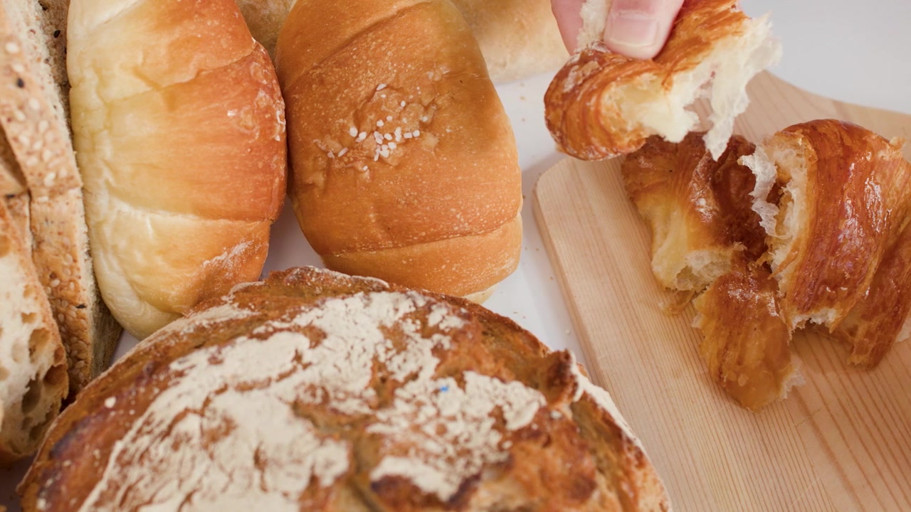 Close-up of hands tearing a flaky croissant on a wooden board with various breads.