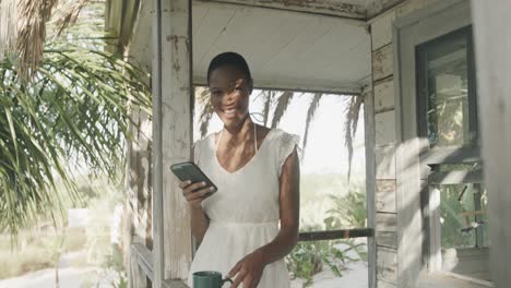 Happy-biracial-woman-using-smartphone-and-drinking-coffee-outside-wooden-beach-house,-slow-motion