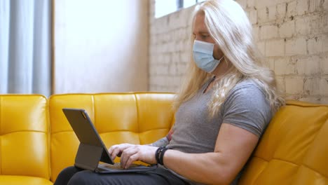 masked long blonde hair bearded caucasian white man, strong muscular and healthy sitting on yellow vegan leather vinyl couch typing on ipad pro magic keyboard - in cinema 4k