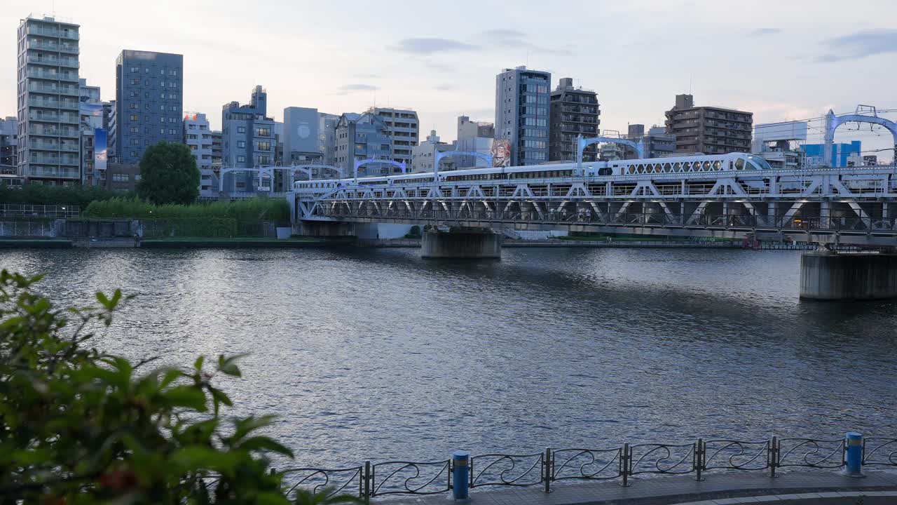 A shot capturing the Tobu Isesaki Line (Tobu Skytree Line) crossing the railway bridge over the Sumida River as it arrives at Asakusa Station. City buildings line the other side of the river