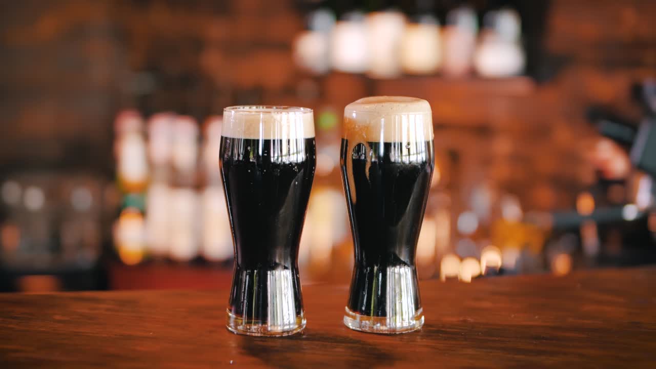 Two glasses of dark stout beer covered with foam, standing on the counter in a pub