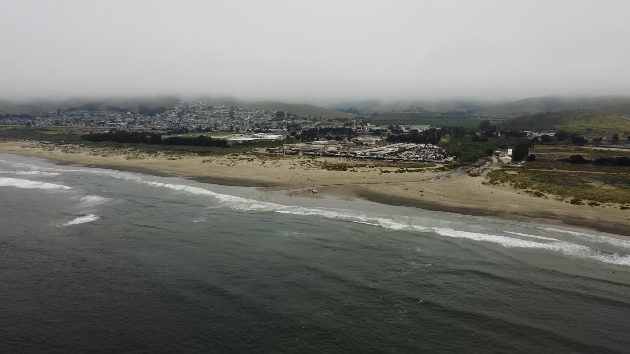 Waves crash onto Morro Beach on the California Coast as fog rolls into the beach town in the background. Aerial shot of this California ton and lush green hills on a cloudy summer day.