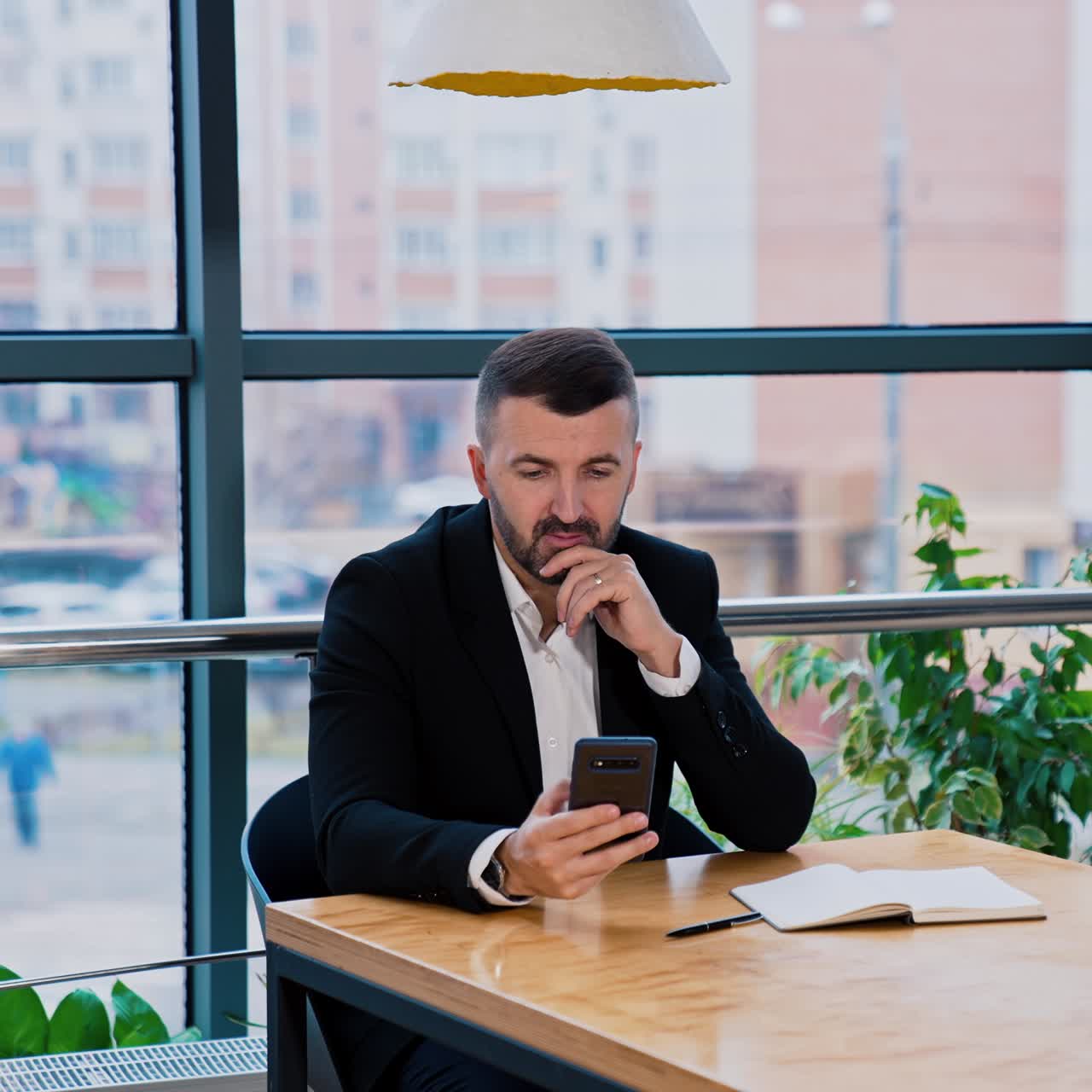 Serious man wearing suit sits at desk talking and holding phone. Boss searching for information in internet on the smartphone