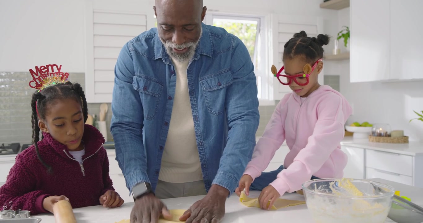 Famiglia afroamericana che prepara dolci per le feste, un anziano guida due bambine a stendere l'impasto sull'isola della cucina