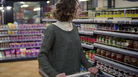 closeup caucasian woman walk with a cart near shop shelves checking a glass jar in grocery market. female customer checking product ingredients. supermarket, sale, shopping, assortment, consumerism concept