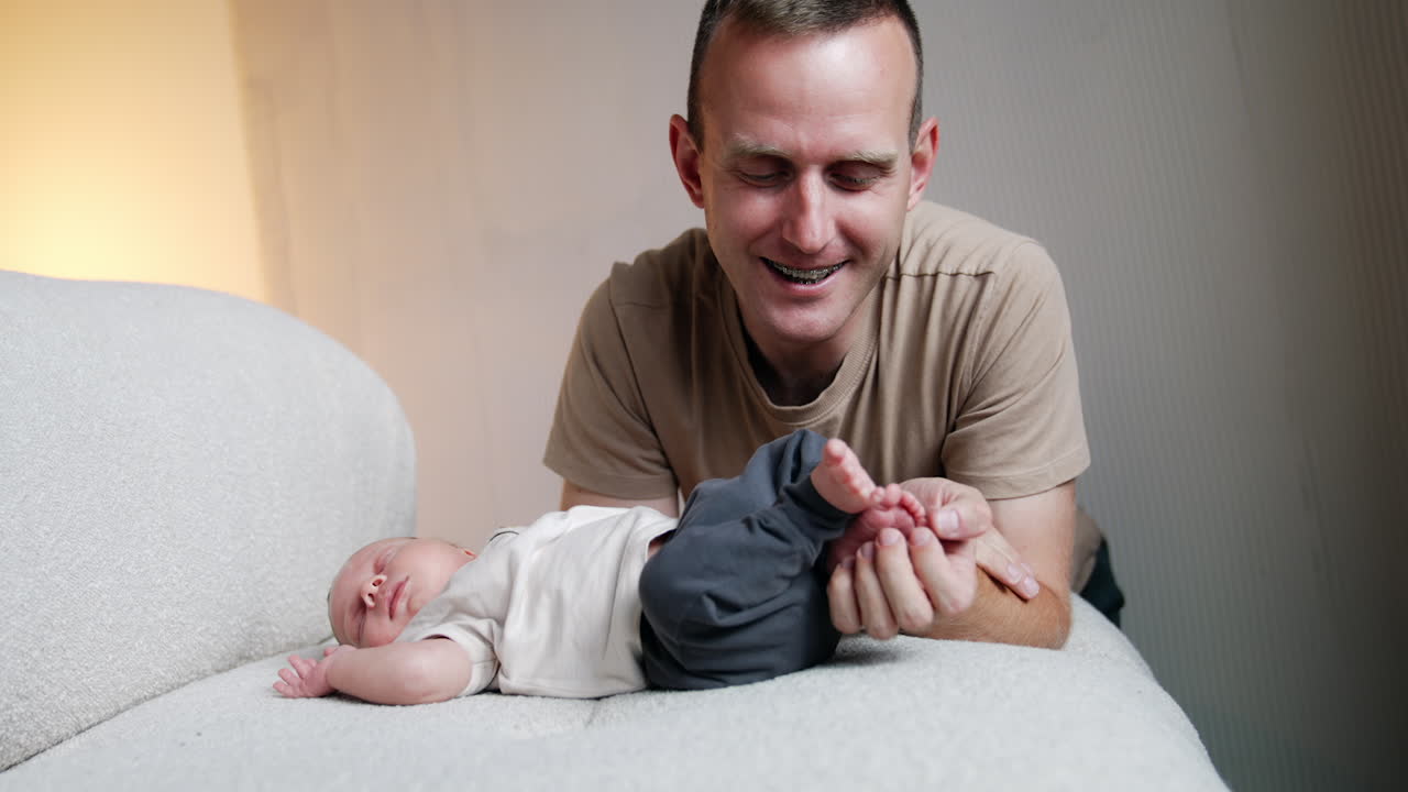 Smiling Caucasian man sits near his sleeping infant son. Father holds and strokes tiny feet of his baby and kisses them.