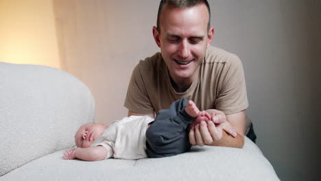 smiling caucasian man sits near his sleeping infant son. father holds and strokes tiny feet of his baby and kisses them.