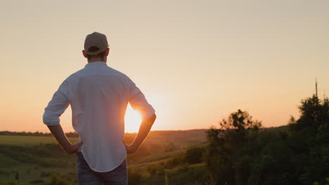 un joven agricultor confiado está de pie frente a un pintoresco campo mientras el sol se pone. vista desde la parte de atrás