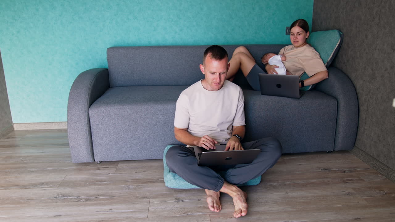 Caucasian parents of a newborn working at home via internet. Woman with a baby sits on sofa and man with laptop sits on the floor beside.