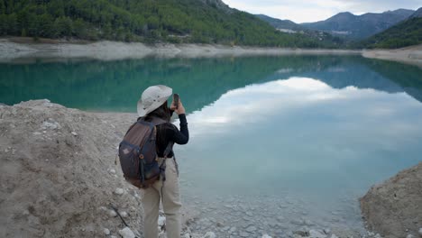 woman hiking and photographing a turquoise lake with mountains