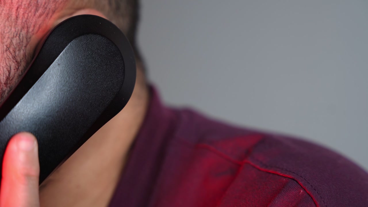髭を生やした男性が黒い固定電話の受話器を耳に当てている超クローズアップショット。スタジオのシーンは、ニュートラルなグレーの背景にドラマチックな赤い照明が特徴的。