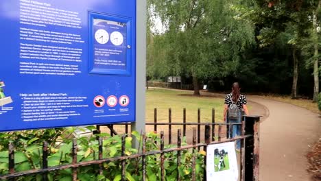 Toma-Panorámica-Del-Cartel-De-Información-De-Holland-Park-Con-Una-Mujer-Joven-Caminando-Por-El-Parque