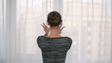 back view of young woman coming up to the window unveiling curtains and looking out of window. enjoying the view outside