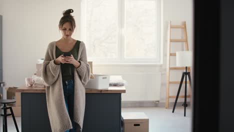 joven mujer caucásica usando un teléfono móvil mientras desempaqueta cosas en una casa nueva.