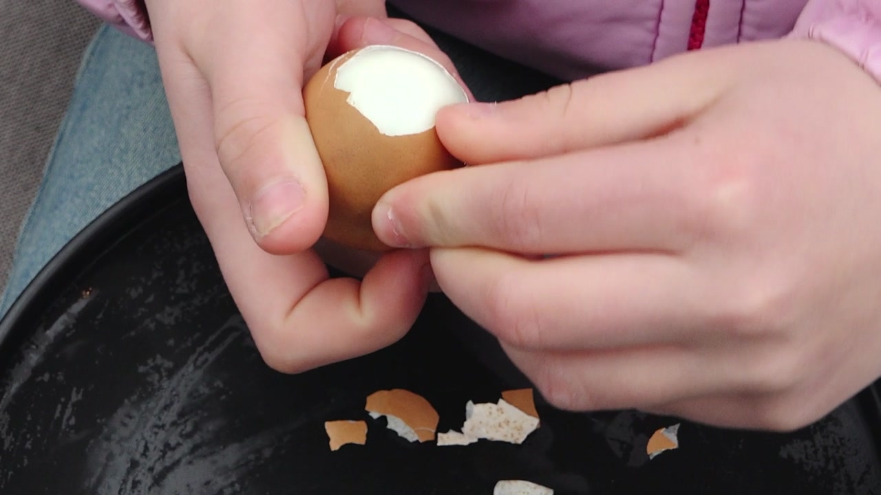 Close-up of a child’s hands peeling a boiled egg over a plate with scattered shells, with steam rising and a casual outdoor setting in the background.