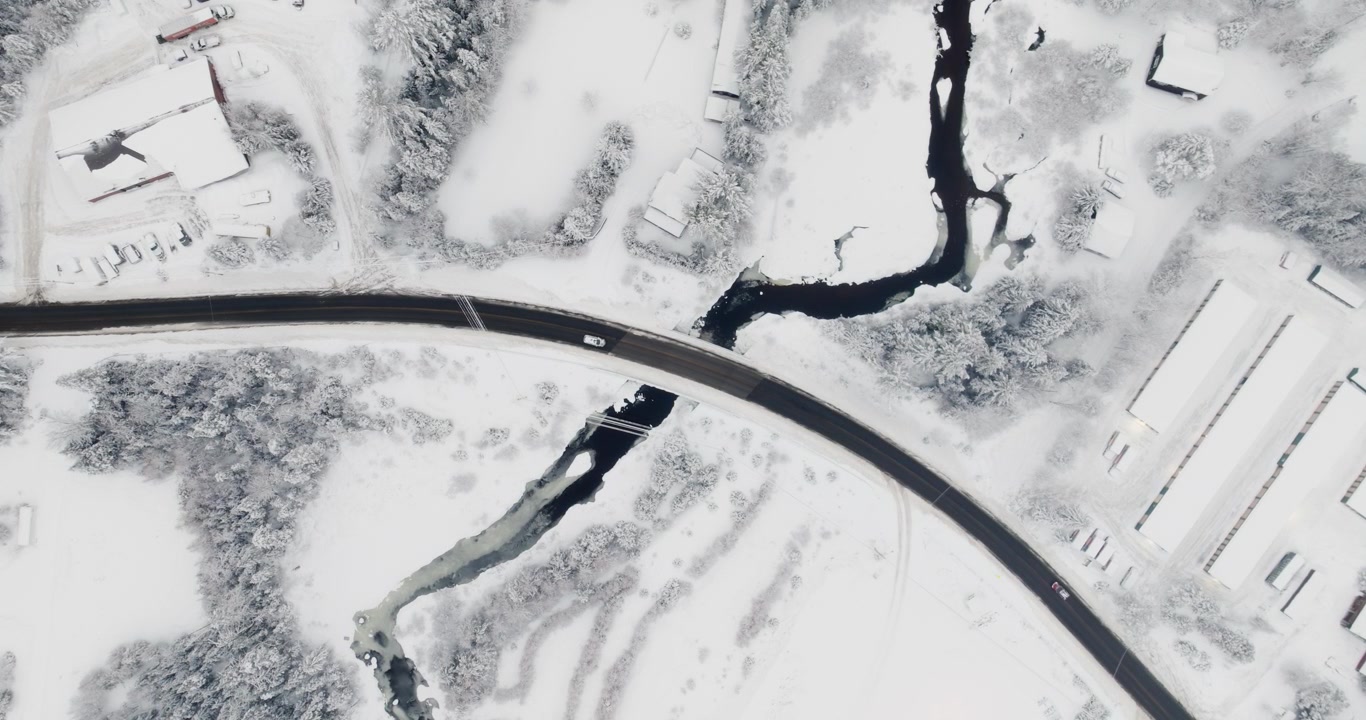 Static top-down drone shot over Lake Placid after fresh snowfall, showcasing a curving road crossing a dark winding river, surrounded by snow-covered terrain and scattered rural structures.