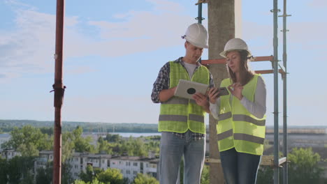 Engineers-builders-man-and-woman-standing-on-the-roof-of-the-building-with-a-tablet-computer-discussing-in-white-helmets-and-shirts.