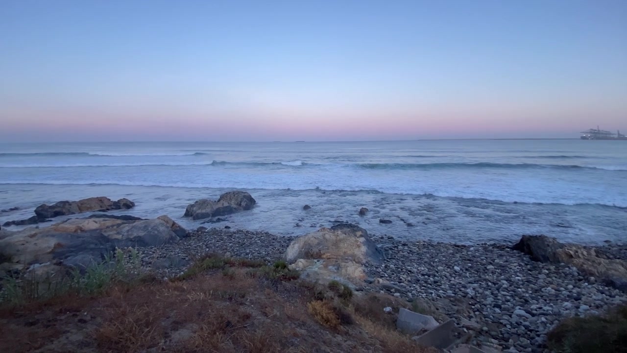 Ondas do oceano quebrando sobre as rochas na costa alentejana, refletindo cores pastel suaves do céu do entardecer.
