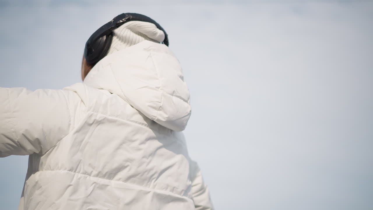 Lower view of elegant young woman spinning around excitedly while wearing over-ear headphones and knit beanie, captured outdoors against pale sky with joyful dynamic energy