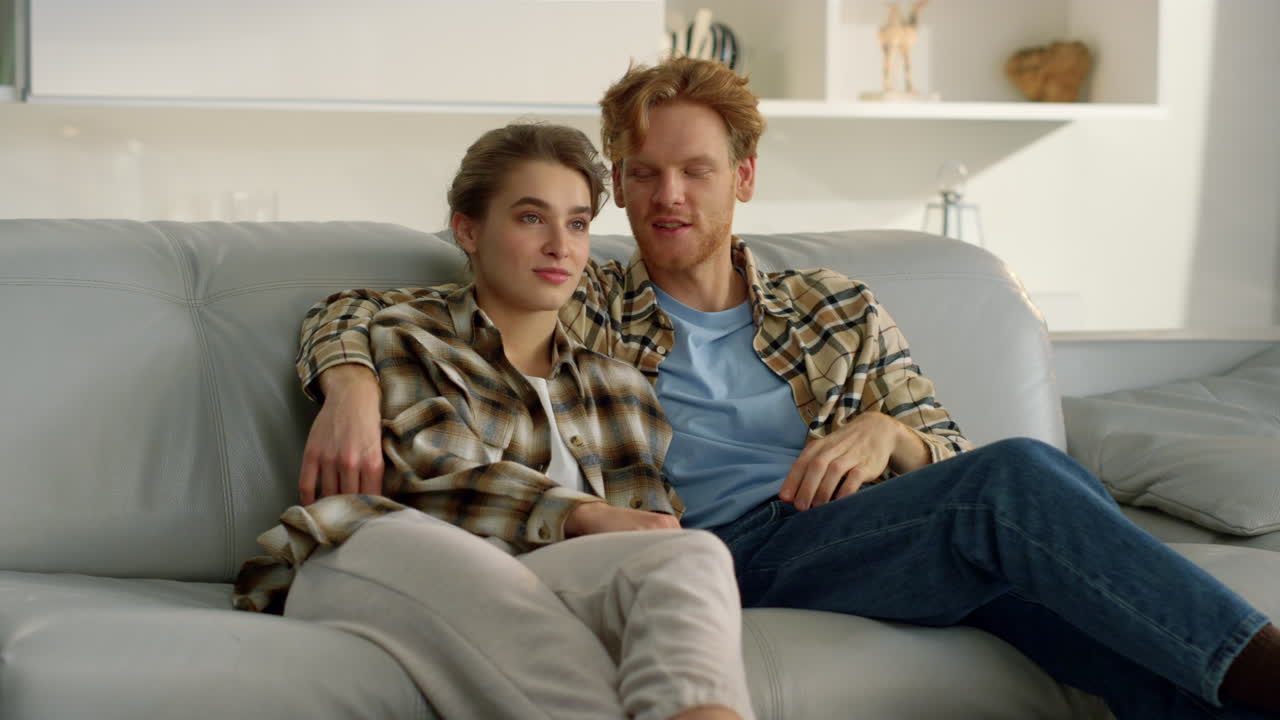 젊은 부부가 집에서 휴식을 취하고 tv를 보고 있다. 웃는 홍색 남자가 리모컨을 사용하고 있다