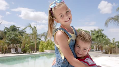 Portrait-of-happy-caucasian-siblings-embracing-at-swimming-pool-at-beach-house