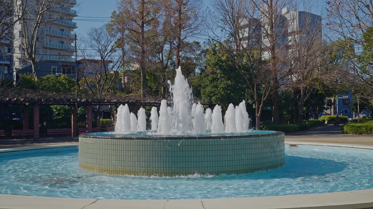 Una toma panorámica de la fuente de agua central rociando en el Parque Gyosen, rodeada de árboles verdes y edificios en un día soleado y brillante.