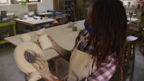 Female-african-american-potter-wearing-face-mask-and-apron-holding-pottery-at-pottery-studio