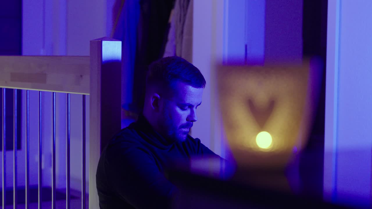 Lonely man sitting at home at night in blue lighting, looking devastated while a glowing heart candle sits in the foreground. Concept for heartbreak, depression, isolation, and emotional crisis