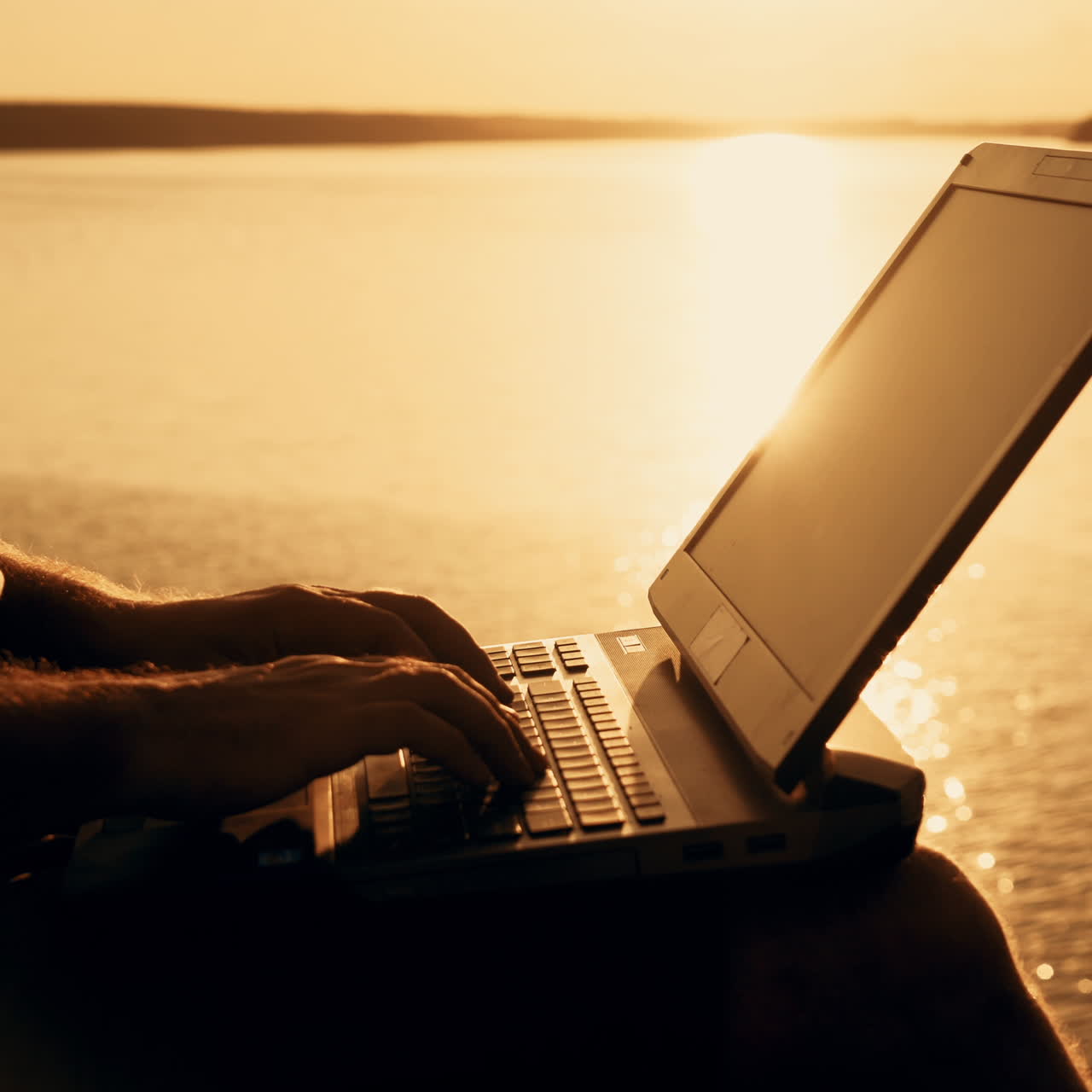A man is sitting and working on the laptop near the lake in the background of the sunset at the weekend. Close-up