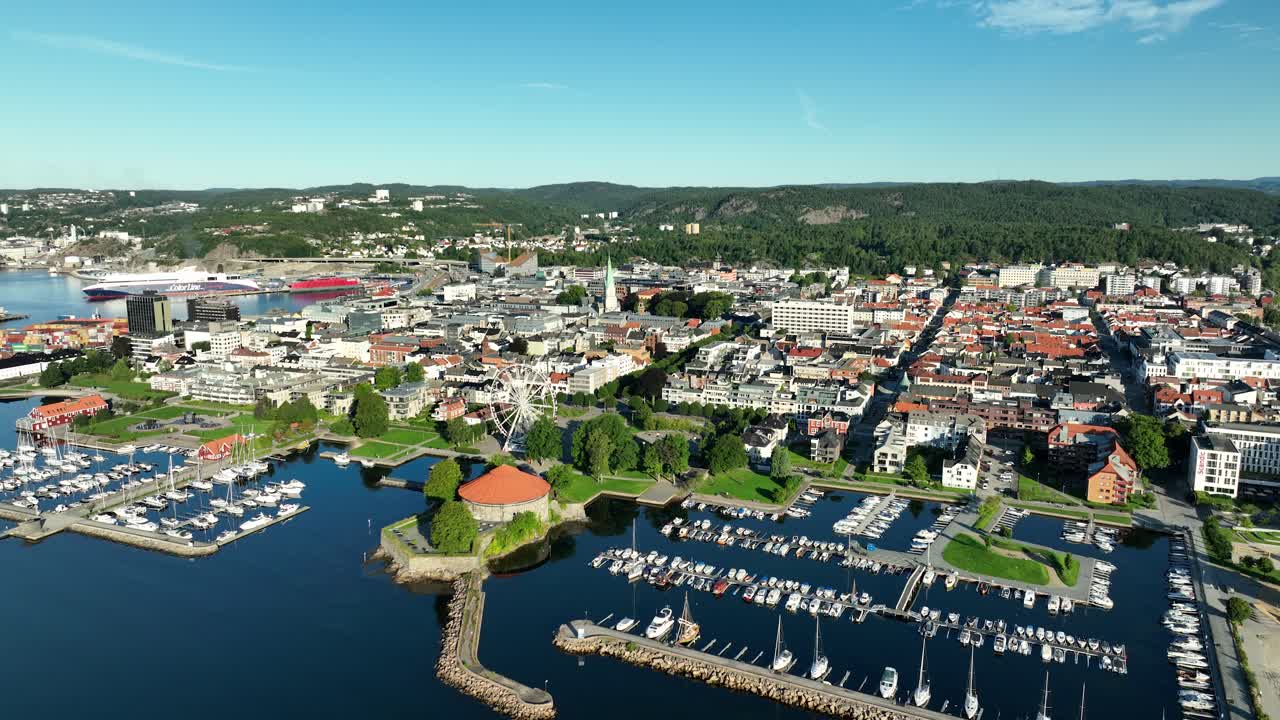 This vibrant coastal town Kristiansand in Norway features a busy marina filled with boats. Colorful buildings line the waterfront, surrounded by lush greenery under a clear blue sky