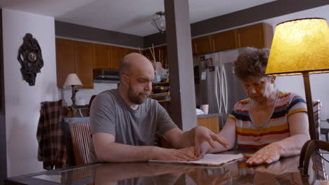 middle-aged male caregiver assists an elderly female stroke survivor by gently stimulating and mobilizing her hemiplegic right thumb during a hand therapy session at the kitchen table, on a cloth pad.