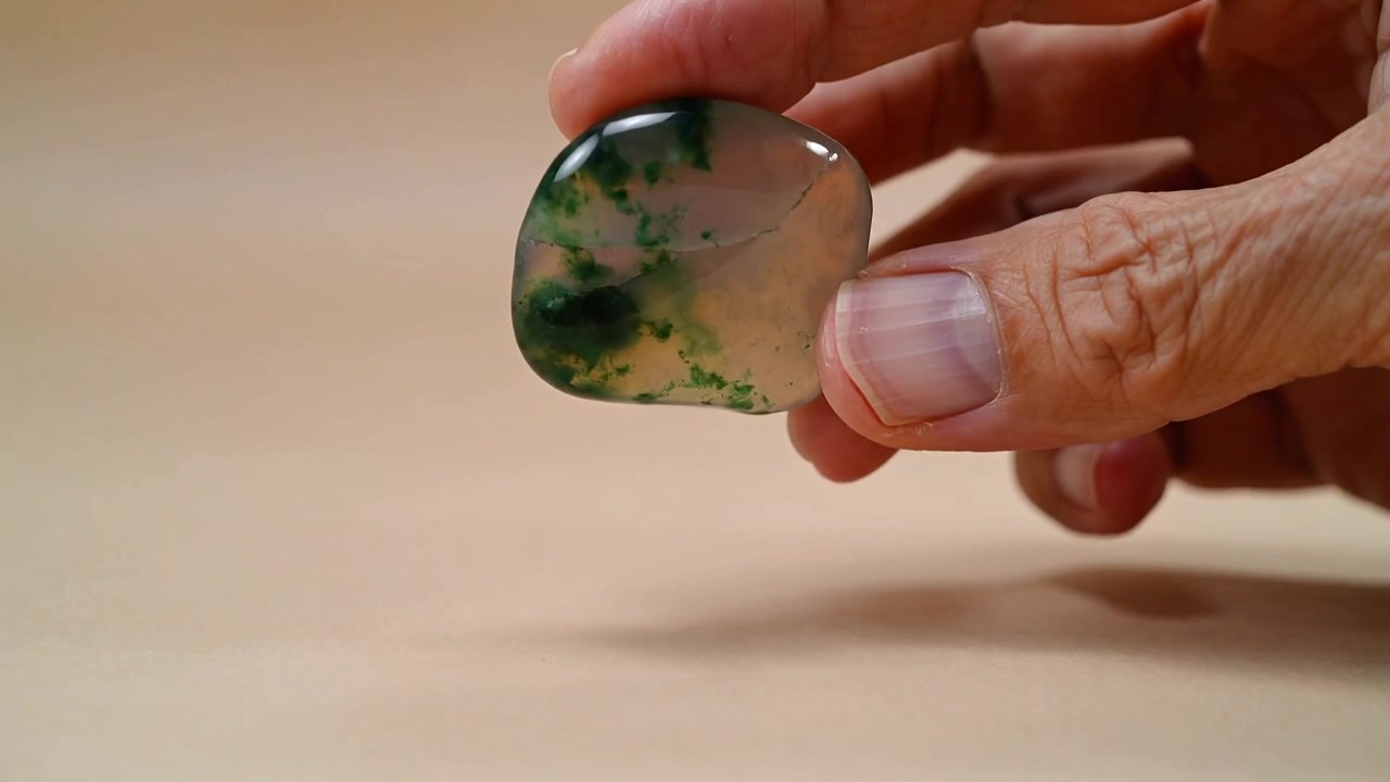 Mano destra che si avvicina, solleva e ruota una pietra lucida su un tavolo di legno, mostrando inclusioni verdi
