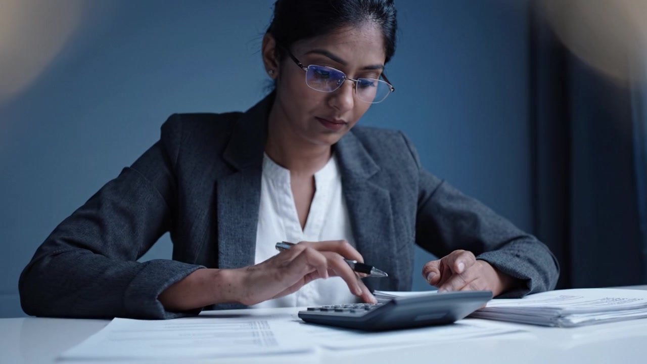 Professionista concentrato calcola dati finanziari con precisione usando una calcolatrice, circondato da documenti e carte organizzati in un ambiente d'ufficio calmo.