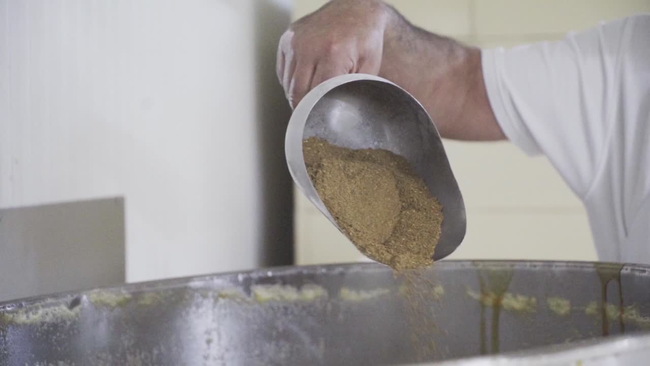 Slow motion handheld shot of a baker using a metal spice shovel to put a mixture of cinnamon into a large mixing pot with a dough for sweets in a sweets factory in Medina Sidonia