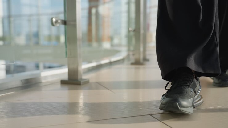 Close up leg view of person wearing black sneakers and wide black pants walking steadily on polished tile floor alongside transparent glass railing in bright modern interior