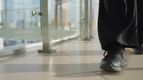close up leg view of person wearing black sneakers and wide black pants walking steadily on polished tile floor alongside transparent glass railing in bright modern interior