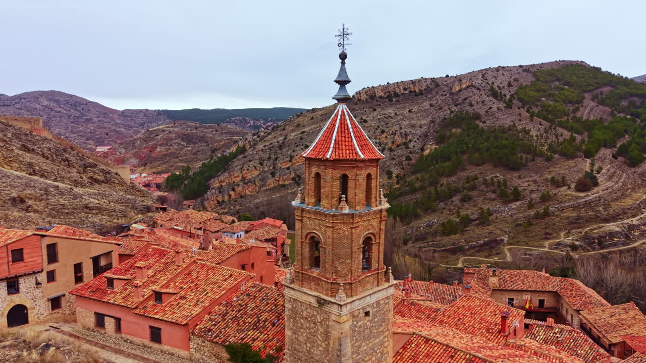 Un'antica torre di chiesa spicca in un villaggio di montagna con tetti rossi e alberi verdi in Spagna, in una giornata nuvolosa.