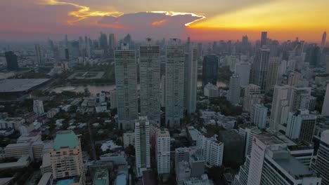 Desde-Una-Perspectiva-Aérea,-Emerge-El-Horizonte-De-Bangkok,-Mostrando-La-Vida-Urbana-Contra-Un-Cielo-Donde-El-Sol-Se-Asoma-A-Través-De-Nubes-De-Tonos-Carmesí,-Creando-Una-Escena-Sorprendente.