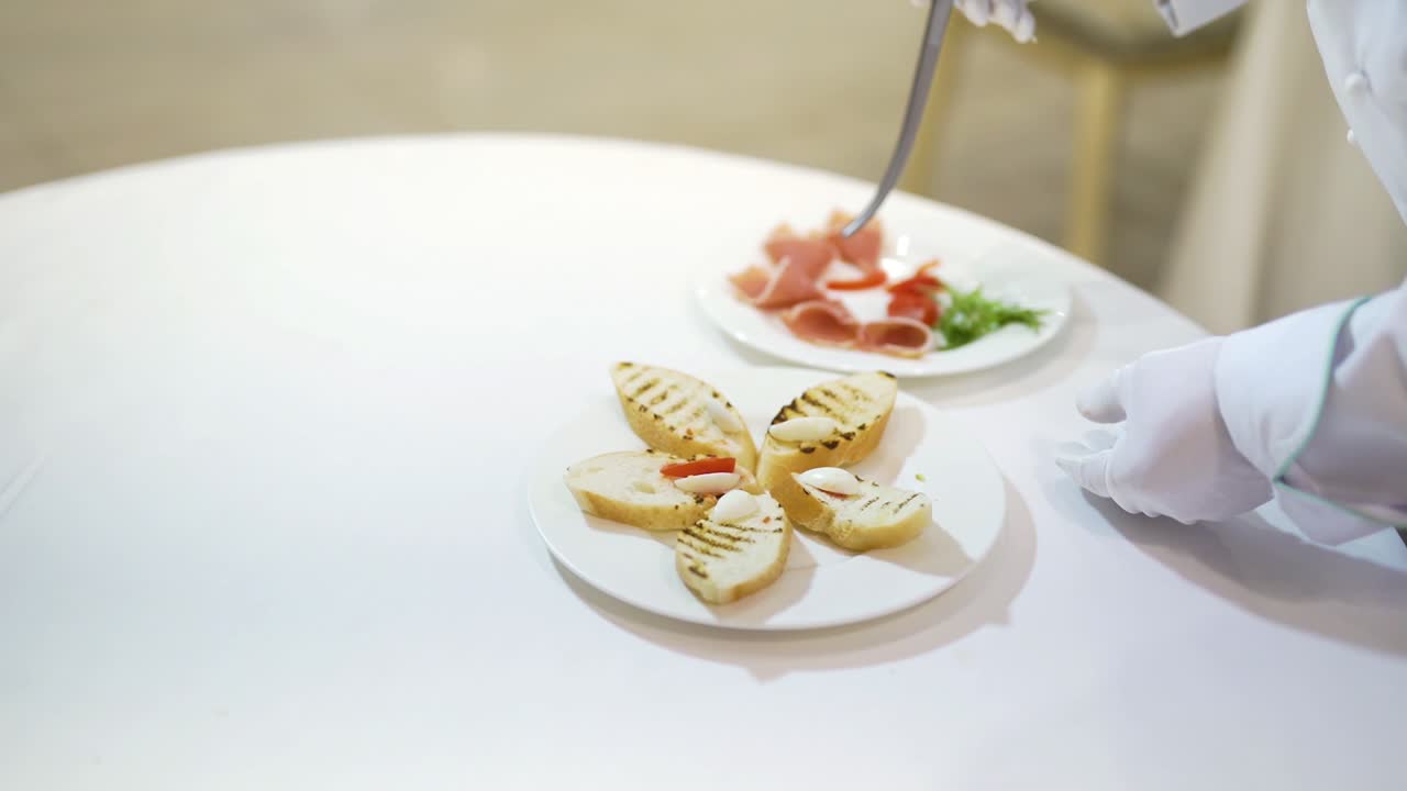 Professional cook decorates toasts with slices of meat, tomatoes by kitchen pinchers. Beautiful presentation of tasty toasts on a white plate for guests